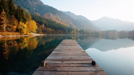 Obraz premium Wooden pier extending into a calm lake with autumn foliage and mountains reflecting on the water surface. Peaceful outdoor landscape vision.