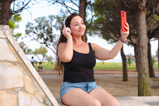 Smiling young woman sitting outdoors wearing headphones and holding red smartphone while taking selfie in park. Modern lifestyle, technology, leisure, freedom, music, summer, youth and urban concept.