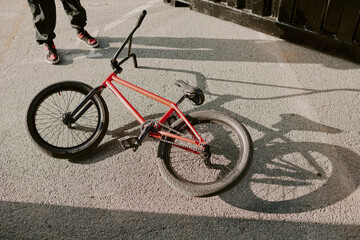 BMX bike lying on pavement casting shadow in sunlight, lower body of teen boy standing nearby wearing sneakers, urban outdoor setting, action paused