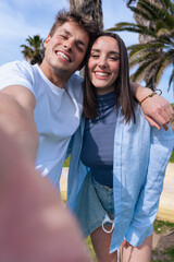 Smiling young couple taking a cheerful selfie together on a sunny day at the beach, with palm trees...