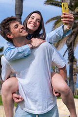 Young happy couple outdoors with palm trees, woman hugging man while taking selfie with yellow...