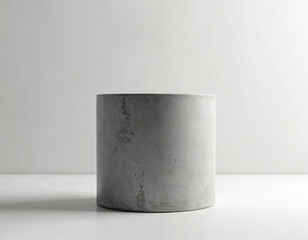 Round grey concrete cylinder pedestal stands on white surface against light wall. Minimalist design object used for product display or architectural element.