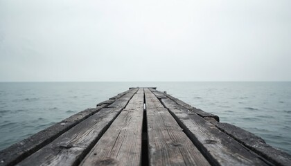 Old wooden pier extends over calm grey sea towards horizon. Empty overcast day at quiet water shoreline. Natural landscape path leads to ocean surface.