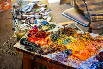 Artist creative workspace with oil paint palette, colorful brushstrokes, paint tubes, glass jar, chair and canvas in the background. Real studio atmosphere full of artistic energy.