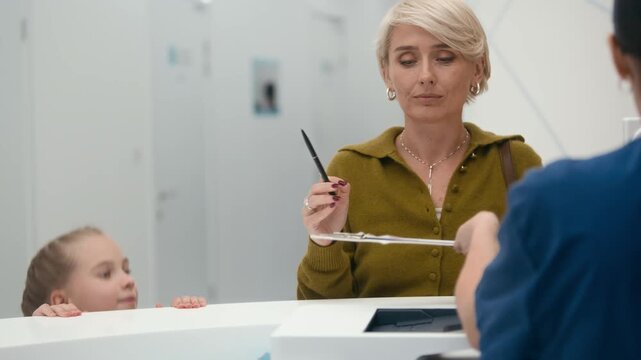 Medium shot of female receptionist in uniform consulting Caucasian woman with daughter and asking to sign medical document in modern clinic