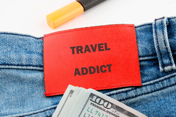 A red tag reading travel addict is attached to a pair of jeans, resting beside a stack of cash. This setup reflects a passion for travel and the excitement of new journeys