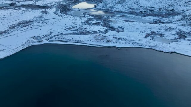 Aerial view of a remote arctic landscape with vast snow-covered hills, multiple frozen lakes, and a row of small domes or tents near the coastline