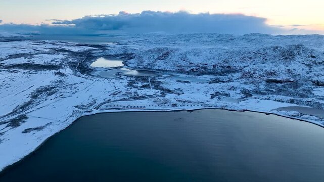 Aerial view of a remote arctic landscape with vast snow-covered hills, multiple frozen lakes, and a row of small domes or tents near the coastline and open water