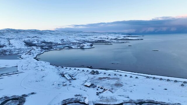 Aerial view of a remote arctic landscape featuring a row of geodesic domes along the coastline next to a cluster of buildings, with vast snow-covered hills and the sea
