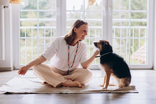 Middle aged woman practices Kundalini yoga at home with her dog nearby. Spiritual balance, feminine energy, harmony, mindfulness, and emotional wellness with animals - Powered by Adobe