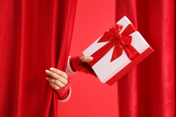 Female hand in Santa costume holding Christmas gift behind curtains on red background