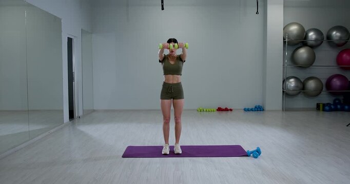 fit woman performs front dumbbell raises showcasing her strength and dedication to fitness while standing on a workout mat. Weights are visible nearby.