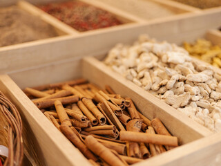 Different types of spices are neatly arranged in wooden boxes, showcasing cinnamon sticks, dried ginger, and various herbs in a bustling market during the afternoon