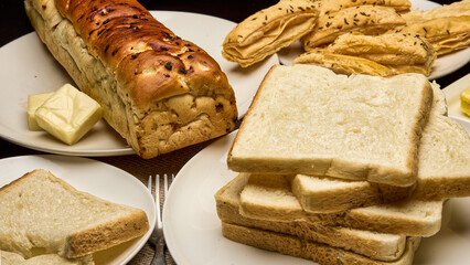 A high-quality close-up photograph of assorted freshly baked bread, white toast slices, and a loaf served with butter on a breakfast table. The image highlights the rich texture, golden crust