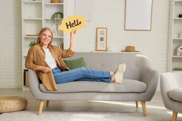 Obraz premium Young woman holding speech bubble with word HELLO on sofa at home