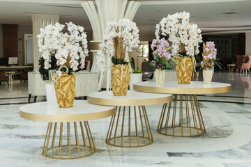 Three stylish tables are displayed in a hotel lobby, adorned with beautiful floral arrangements in decorative vases. The setting features modern decor and bright lighting