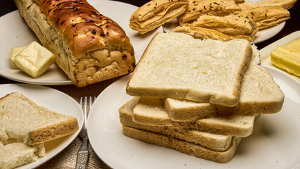 A detailed close-up photo of freshly baked white bread slices stacked neatly beside a golden-brown loaf, puff pastries, and butter cubes on a breakfast table. Perfect image for bakery advertisements