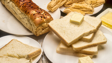 A high-resolution close-up image showing fresh white bread slices stacked on a plate with a cube of butter on top, accompanied by a golden-brown loaf and crisp puff pastries in the background