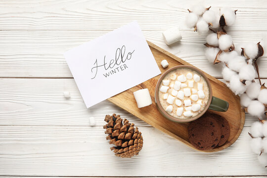 Paper with text HELLO WINTER, cocoa, marshmallows and cotton flowers on white wooden background