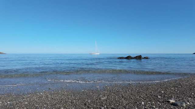 Waves gently lap on the shore of Portnadler Bay in Cornwall, near Looe and Talland. A sail boat is is anchored in the cove. Beautiful sunny day. Shingle beach. 