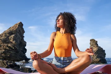 Fotobehang Lotusbloem Young woman meditating in lotus pose on a yoga mat by the ocean, feeling serene and connected to nature  © Javier