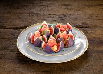 Fresh figs are artistically arranged on a white plate. Each fig is cut and stuffed with cream and ham, garnished with herbs, offering a colorful and appetizing presentation on a wooden table