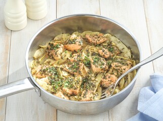 In a pan, tender salmon with herbs is mixed with artichoke hearts, ready to serve. The pan sits on a white wood table next to bottles
