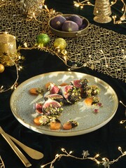 Fresh figs and dried apricots drizzled in chocolate and sprinkled with pistachios are arranged on a plate. Warm lighting and gold decorations create a celebratory atmosphere