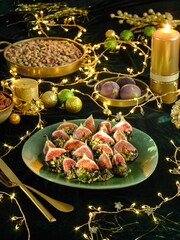 A holiday table is set with a plate of figs dipped in chocolate and pistachio, along with nuts, candle lights, and ornamental balls, creating a warm festive ambiance