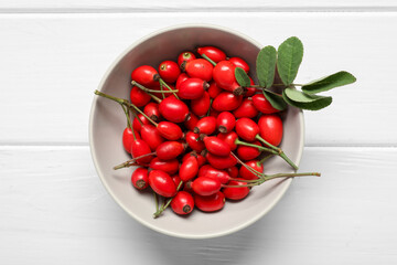 Bowl with fresh rose hip berries and leaves on white wooden background