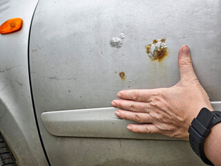 Hand inspecting rust spots on a silver car door near a residential area during daylight