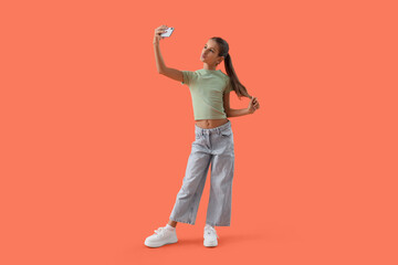 Dreamy teenage girl with mobile phone taking selfie on orange background