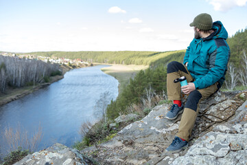 A bearded man is sitting resting on a rock with a thermocup in his hand, a knitted hat on his head