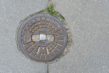 Round metal manhole cover embedded in concrete pavement, slightly weathered with a patch of green grass nearby.