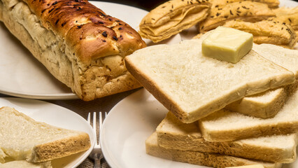 A detailed close-up image of a freshly baked golden-brown bread loaf with soft white bread slices stacked neatly on a plate, topped with a cube of butter. Crispy puff pastries in the background