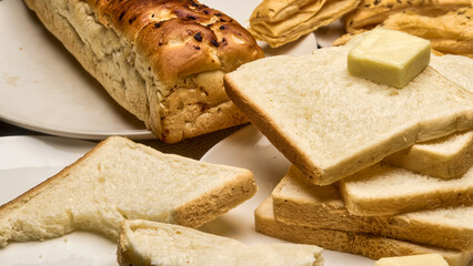 A detailed close-up image of a freshly baked golden-brown bread loaf with soft white bread slices stacked on a plate, topped with a cube of butter. Crisp puff pastries in the background