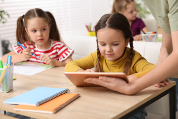 Fototapeta premium Teacher and little girl with tablet at wooden desk in elementary school, closeup