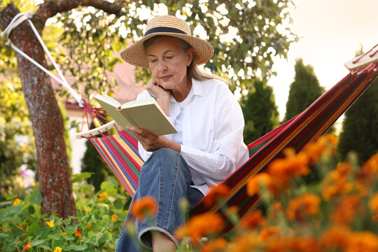 Senior woman with straw hat and book resting in hammock outdoors - Powered by Adobe