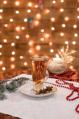 Transparent glass with steaming golden beverage stands on saucer, decorated with cinnamon, star anise, and apple slice. Cozy Christmas lights and pine branch create a holiday atmosphere