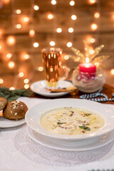 Cozy winter soup with creamy texture and herbs, placed on white tablecloth beside buns and tea glass, glowing golden lights in background.