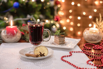 Glass mug with red mulled wine placed beside layered dessert, red beads, burning candles and Christmas lights creating cozy holiday atmosphere.
