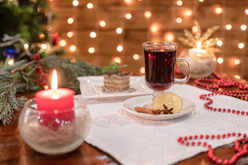 Warm festive still life with glass of mulled wine, glowing red candle, small dessert and Christmas garland creating cozy winter mood.