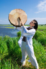 Female shaman in the white dress drumming in the natural environment