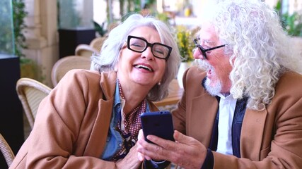 Happy senior couple sitting together at outdoor cafe using smartphone and looking at social media. Elderly people enjoying technology, connection, communication, digital lifestyle, love and leisure - Powered by Adobe