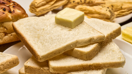 A close-up image of soft, freshly baked white bread slices stacked neatly on a plate with a cube of butter on top. In the background, a golden-brown bread loaf and crispy puff pastries.