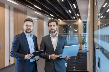 Two professional businessmen holding a clipboard and a laptop, standing in a brightly lit corporate...