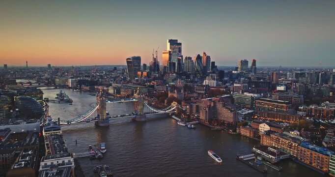 UK, London cityscape at sunset, iconic Tower Bridge spanning the River Thames, modern skyscrapers reflecting golden light against the twilight sky. Travel destination. Aerial drone flight panorama