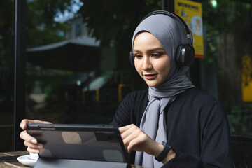 Smiling Muslim women in hijab enjoying coffee and music with headphones while working on a tablet...