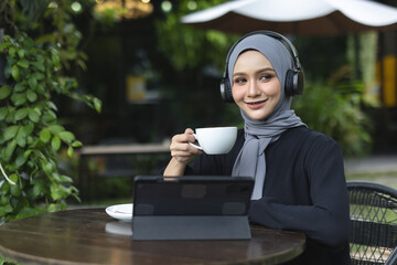 Smiling Muslim women in hijab enjoying coffee and music with headphones while working on a tablet...