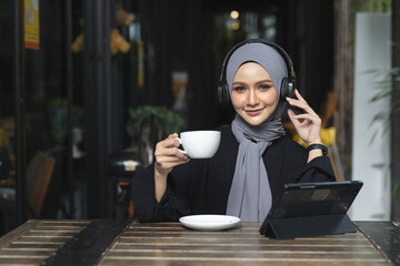 Smiling Muslim women in hijab enjoying coffee and music with headphones while working on a tablet...